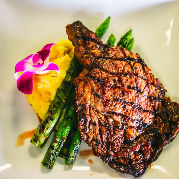 Grilled steak with asparagus, mashed potatoes, and a purple orchid garnish on a white plate.